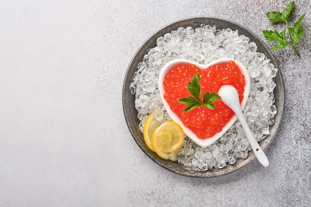 Caviar rojo en un tazón en forma de corazón servido con limón y cubitos de hielo en la mesa de fondo gris.