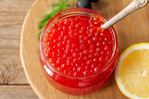 Caviar de trucha salmón rojo en un frasco de vidrio, limón y eneldo sobre fondo de madera.