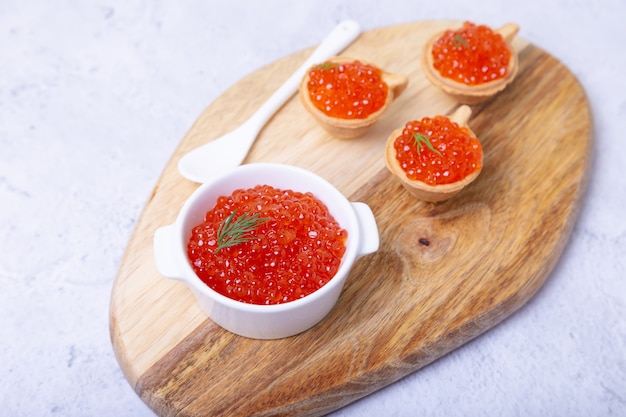 Caviar vermelho (caviar de salmão) em uma tigela branca em uma placa de madeira. No fundo são tortinhas com caviar. Foco seletivo, close-up.