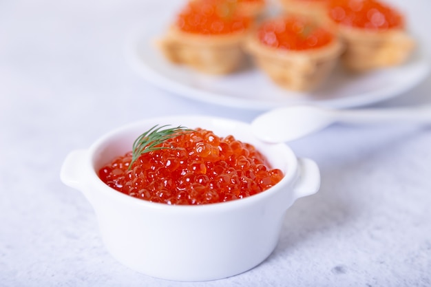 Caviar vermelho (caviar de salmão) em uma xícara branca. Ao fundo, um prato com tortinhas e caviar. Foco seletivo, close-up.