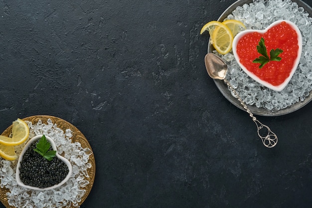 Caviar vermelho e preto em uma tigela em forma de coração servido com limão e cubos de gelo na mesa de fundo preto