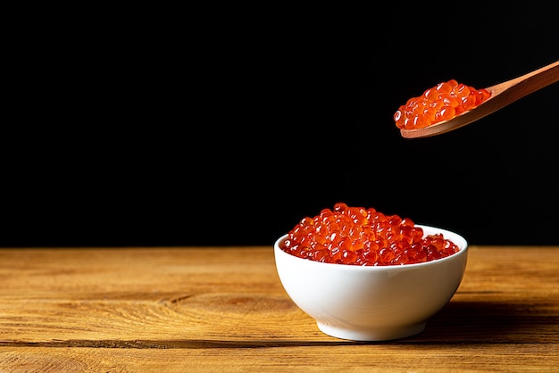 Caviar vermelho em um copo de madeira em um fundo preto com uma colher. Uma grande pilha de caviar brilhante. Caviar fresco e delicioso. Um lugar para publicidade, logotipo, etiqueta, layout, maquete. Layout, modelo.