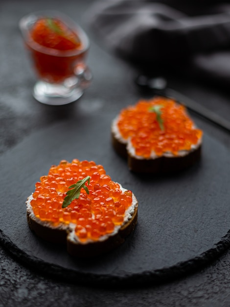 Foto caviar vermelho em uma tigela e sanduíches de caviar em uma placa de pedra preta. em um fundo estrutural preto