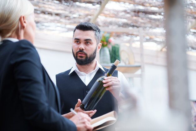 Cavist joven confiado con una botella de vino hablando con su colega durante la discusión de sus características en el trabajo