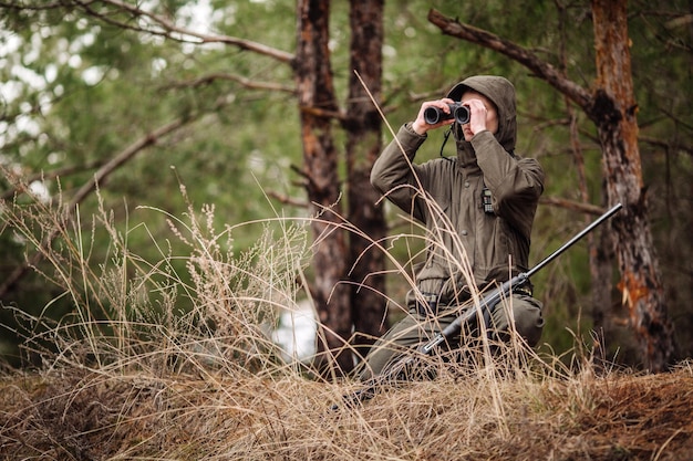 Cazador masculino con binoculares listo para cazar sosteniendo un arma y caminando en el bosque