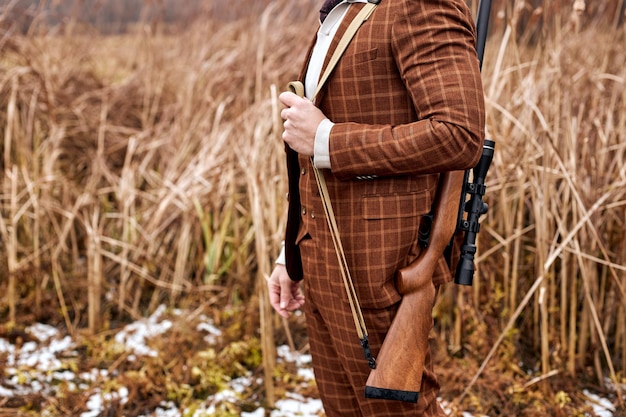 Cazador con rifle se encuentra en la naturaleza busca juego, en la temporada de otoño. Joven varón caucásico en traje marrón de moda, en campo, listo para disparar, esperando animal salvaje en el horizonte