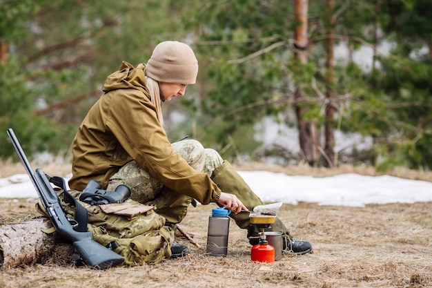 Cazadora preparando comida con un quemador de gas portátil en un bosque de invierno