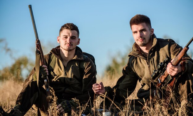 Cazadores con rifles relajándose en el entorno natural. Los cazadores satisfechos con la bebida que calienta la captura. Los amigos de los cazadores disfrutan del ocio. Descanso para el concepto de hombres reales. Caza con amigos hobby ocio.