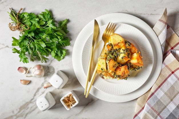 Cazuela de desayuno italiano o estratos de espinacas, queso y pan en cubos remojado durante la noche horneado con mostaza en un plato blanco con cubiertos dorados sobre un fondo de mármol blanco, vista superior de cerca