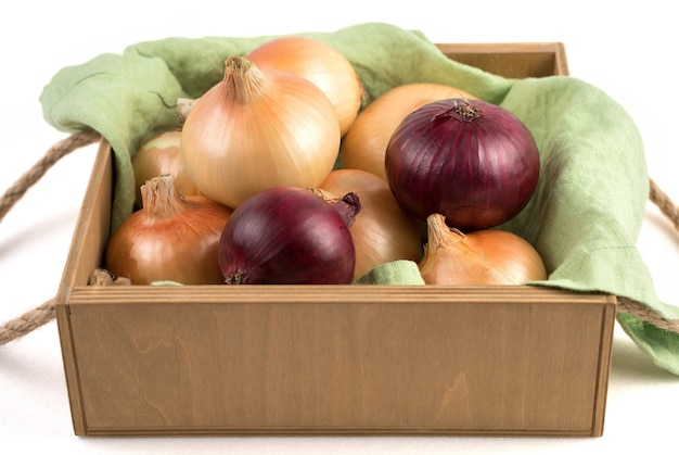 Cebollas de diferentes variedades en una hermosa caja de embalaje de madera sobre una servilleta de lino, aislado. Concepto de productos naturales.