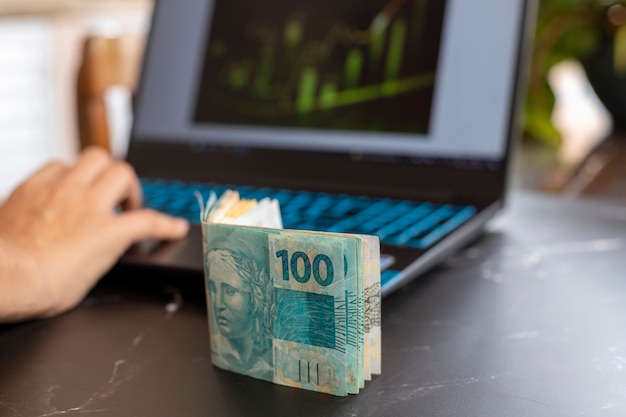 Foto cédulas de dinheiro brasileiro sobre a mesa, com fundo desfocado de laptop