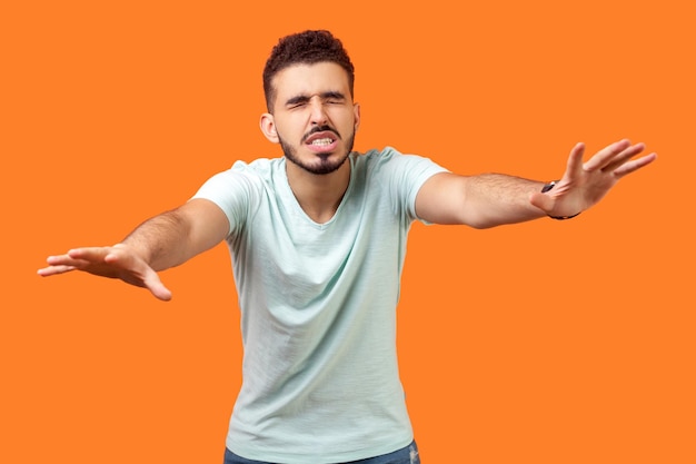 Ceguera. Retrato de un moreno desorientado con barba en una camiseta blanca informal caminando con los ojos cerrados y extendiendo las manos para encontrar el camino. tiro de estudio interior aislado sobre fondo naranja