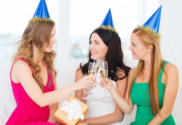 celebração, bebidas, amigos, despedida de solteira, conceito de aniversário - três mulheres sorridentes usando chapéus azuis com taças de champanhe e um presente