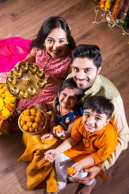 Celebração de Diwali ou Rakshabandhan - jovem família indiana de quatro pessoas celebrando o festival Deepavali ou bhai Dooj com doce laddoo, lâmpada a óleo ou diya e caixas de presente, comendo comida ou tomando selfie