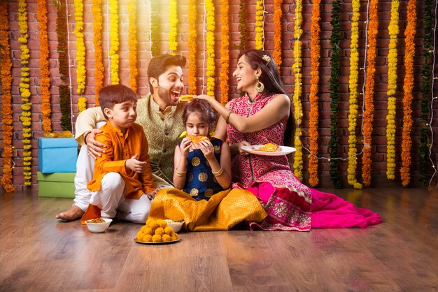 Celebração de Diwali ou Rakshabandhan - jovem família indiana de quatro pessoas celebrando o festival Deepavali ou bhai Dooj com doce laddoo, lâmpada a óleo ou diya e caixas de presente, comendo comida ou tomando selfie