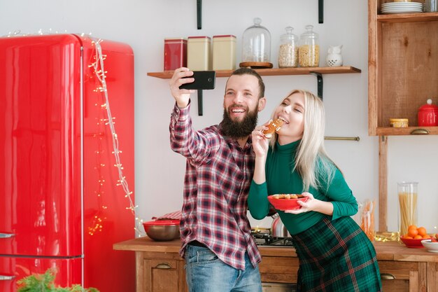 Celebração de natal divertida em casa. casal feliz usando telefone para tirar selfie, comendo biscoitos de gengibre.