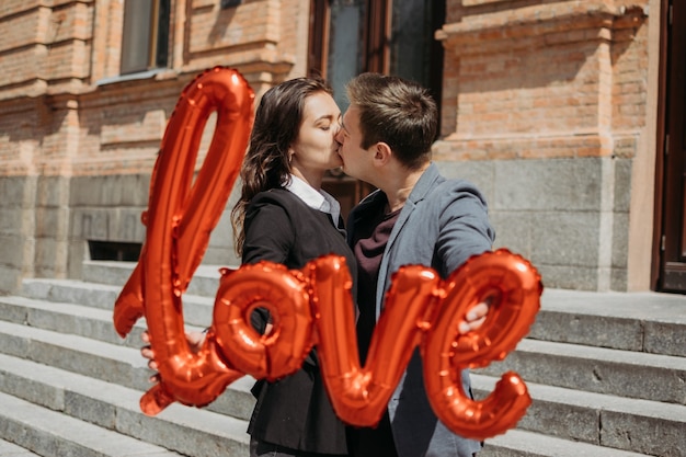 Celebração do dia dos namorados e conceito de namoro. casal amoroso feliz com balões de amor vermelhos na rua da cidade. retrato ao ar livre do jovem casal com balão em forma da palavra amor.