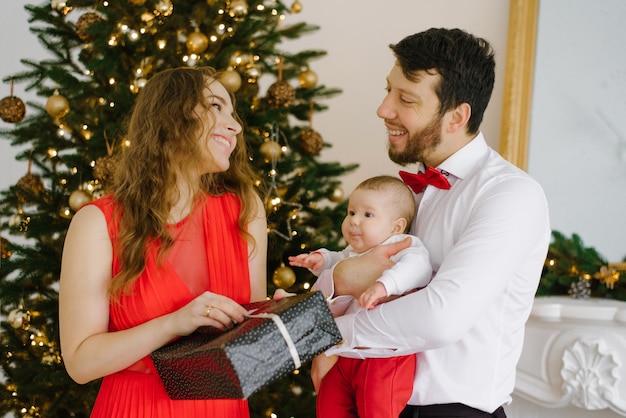 Celebração familiar de Natal e ano novo por uma jovem família com uma criança pequena perto da árvore de Natal. Eles se dão presentes