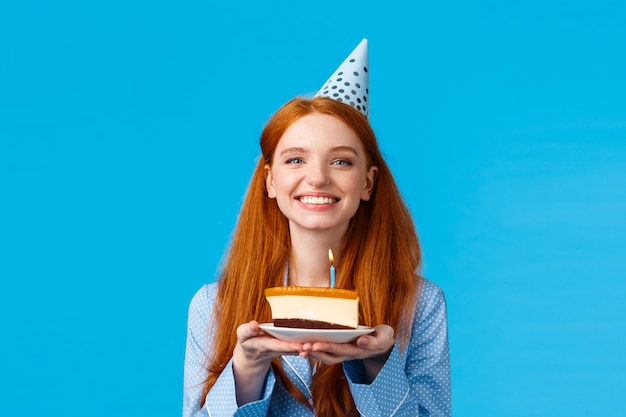 Celebração, feriados e conceito de felicidade. Mulher ruiva concurso e feminina, adorável alegre no boné de b-dia, segurando o pedaço de bolo comemorando aniversário, apagar a vela e fazer desejo