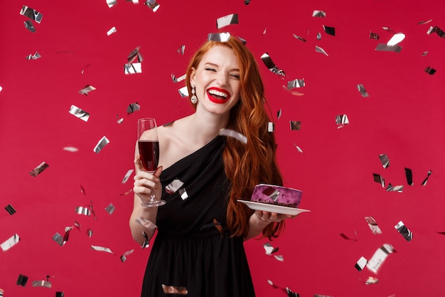 Celebração, feriados e conceito de festa. Retrato de mulher ruiva jovem bonita despreocupada se divertindo no aniversário, comemorando a rir enquanto confetes voando por aí, segure o copo de champanhe e bolo