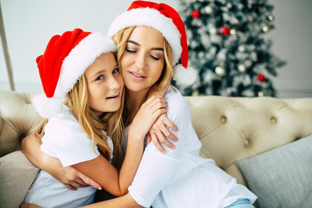 Celebração, feriados, família, natal, ano novo! linda mãe feliz e sua filhinha de chapéu de papai noel está sentada e abraçando uma à outra no fundo da árvore de natal