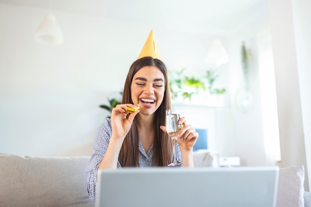 Celebração virtual. mulher feliz com chapéu de festa comemorando aniversário online em quarentena ou auto-isolamento, usando laptop para videochamada com amigos e familiares, segurando champanhe
