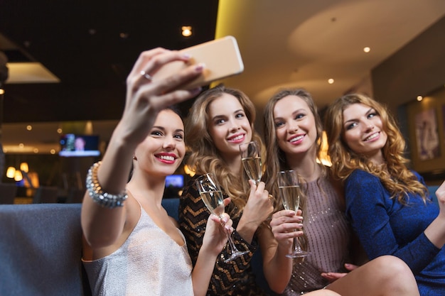 celebración, amigos, despedida de soltera, tecnología y concepto de vacaciones - mujeres felices con copas de champán y smartphone tomando selfie en el club nocturno