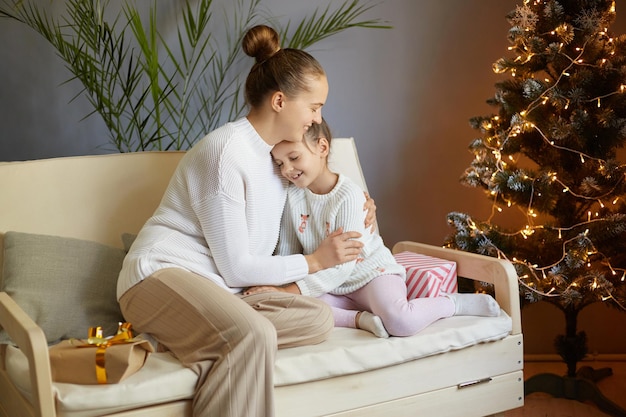 Celebración de Año Nuevo en casa Retrato de una joven madre adulta sentada con su hija en casa cerca del árbol de Navidad y disfrutando de una cálida y acogedora noche de Navidad mamá abrazando a su amado hijo