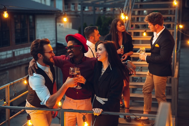 Celebración corporativa a la cálida luz de las lámparas en la noche de verano, jóvenes amigos, colegas se ven felices, conversan, se divierten. Vacaciones, fines de semana, negocios y finanzas, concepto de amistad. Teambuilding.