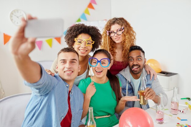 Foto celebración corporativa y concepto de vacaciones amigos o equipo felices con accesorios de fiesta y bebidas no alcohólicas tomando selfies en la oficina