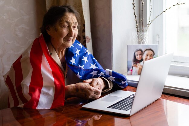 Celebración del Día de la Independencia después de la cuarentena. Granny mirando la pantalla de un portátil y celebrando la fiesta nacional con su familia en línea, se preocupa por las personas mayores, la tecnología personal, se centra en la mujer.