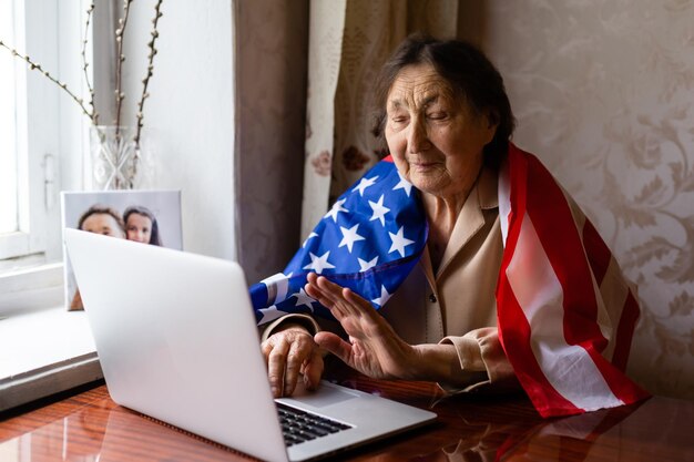 Celebración del Día de la Independencia después de la cuarentena. Granny mirando la pantalla de un portátil y celebrando la fiesta nacional con su familia en línea, se preocupa por las personas mayores, la tecnología personal, se centra en la mujer.