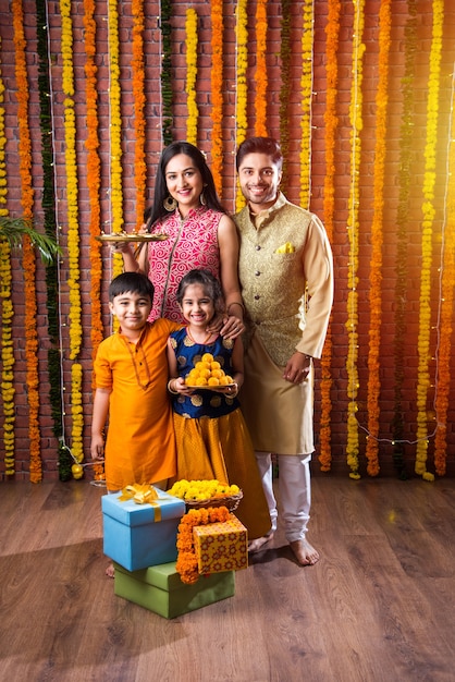 Celebración de Diwali o Rakshabandhan: joven familia india de cuatro miembros que celebra el festival Deepavali o bhai Dooj con laddoo dulce, lámpara de aceite o diya y cajas de regalo, comiendo o tomando selfie