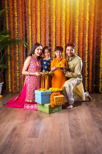 Celebración de Diwali o Rakshabandhan: joven familia india de cuatro miembros que celebra el festival Deepavali o bhai Dooj con laddoo dulce, lámpara de aceite o diya y cajas de regalo, comiendo o tomando selfie