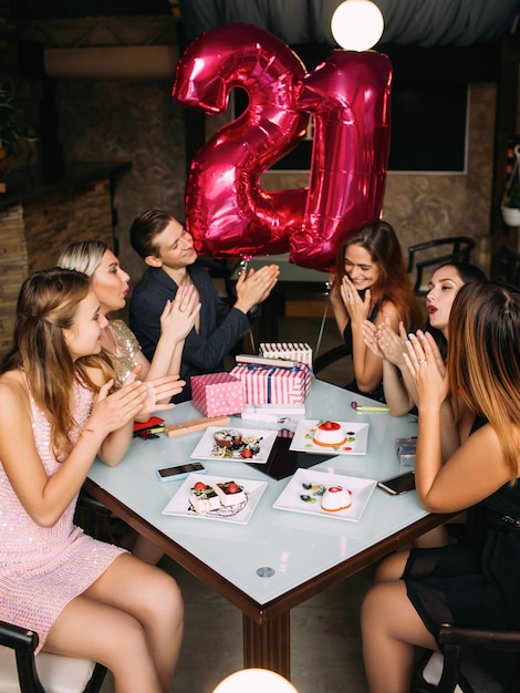 Celebración de la fiesta de cumpleaños número 21 de chica. Compañía de amigos felicitando a la mujer joven en el café, la vida juvenil moderna. Estado de ánimo alegre, concepto de edad adulta