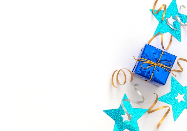 Celebración de fondo blanco con caja de regalo con cinta dorada, serpentinas de fiesta brillantes y estrellas azules.