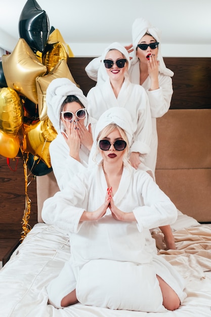 Celebración en el spa. Diversión y relajación de fiesta. Mujeres sonrientes posando en la cama. Gafas de sol, albornoces y turbantes de toalla puestos.
