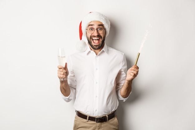 Celebración y vacaciones de invierno. Chico feliz con sombrero de Santa regocijándose en la fiesta de año nuevo, bebiendo champán y gritando de alegría, fondo blanco.