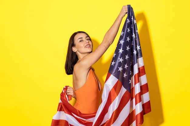 Celebración del verano y el patriotismo Una joven bronceada por el sol se ve sexy sin esfuerzo en su traje de baño mientras sostiene la bandera estadounidense sobre un fondo amarillo