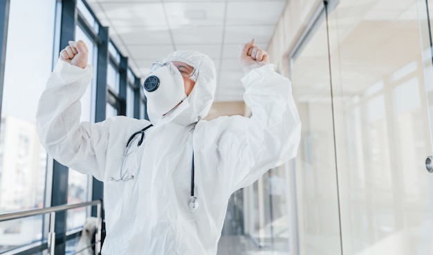Celebrando a vitória. Cientista médica no jaleco, óculos de proteção e máscara em pé dentro de casa