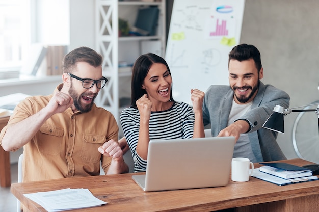 Foto celebrando el éxito. tres hombres de negocios felices en ropa casual elegante mirando la computadora portátil y gesticulando