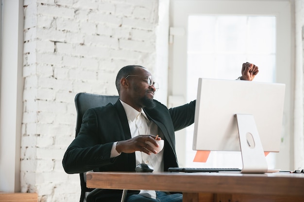 Celebrando la victoria. Emprendedor afroamericano, empresario que trabaja concentrado en la oficina. Se ve feliz, alegre, con traje clásico, chaqueta. Concepto de trabajo, finanzas, negocios, liderazgo de éxito.