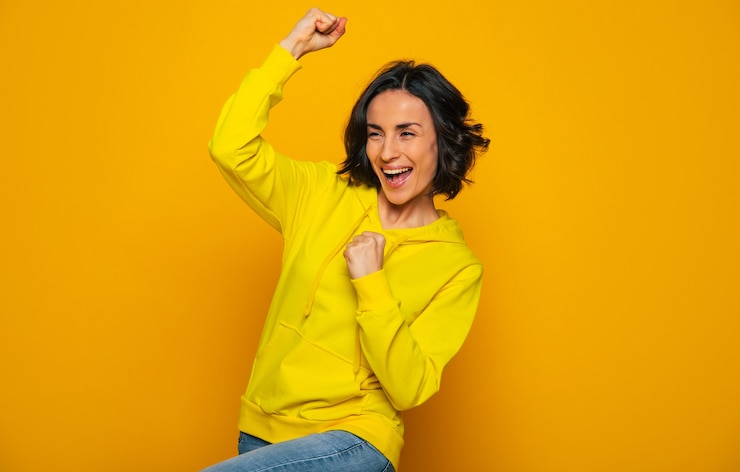 Foto celebrando la victoria. sonriente niña feliz vestida con una sudadera con capucha amarilla, celebrando la victoria con los puños cerrados, mirando hacia los lados.