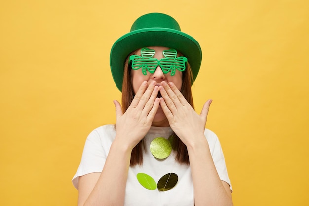 Celebrar las tradiciones irlandesas diversión y vacaciones festivas extremadamente sorprendida mujer con sombrero verde festivo y gafas de trébol de pie aislado sobre fondo amarillo ve algo increíble