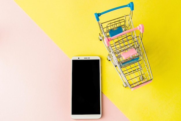 Celular branco de tela em branco e carrinhos de compras em fundo rosa pastel e amarelo Estilo minimalista flatlay