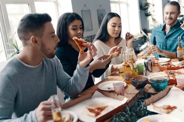 Cena con amigos. Grupo de jóvenes en ropa casual comiendo pizza y sonriendo mientras tienen una cena en el interior