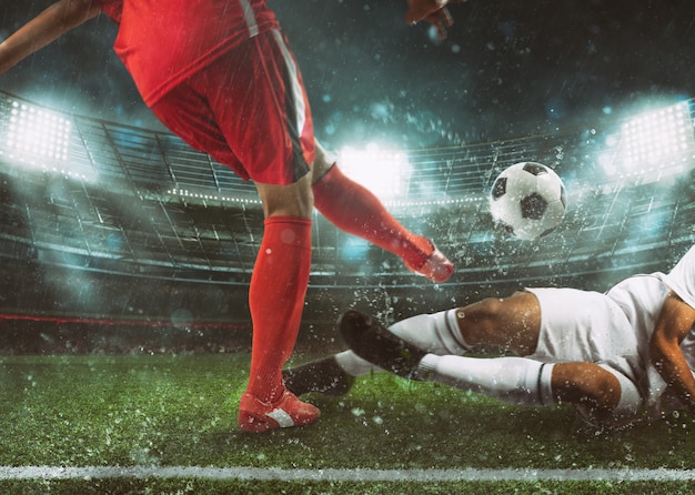 Cena de futebol no estádio com jogador de uniforme vermelho chutando a bola e adversário em tackle para defender