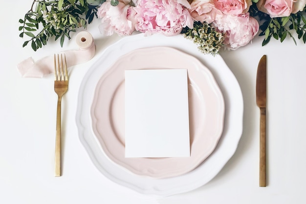 Cena de maquete de mesa de aniversário de casamento feminino Pratos de porcelana papel em branco cartão de menu fita folha de talheres douradorosas flores de peônia Fundo de mesa branca Vista superior plana leiga