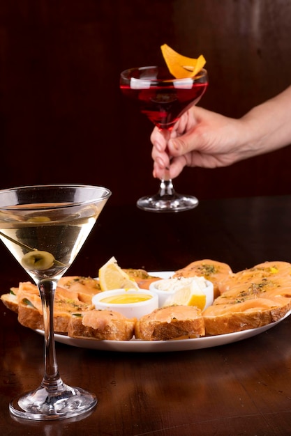 Foto cena con plato de bruschetta de salmón ahumado con pepino y queso mozzarella mano femenina recogiendo copa de cóctel sobre fondo borroso