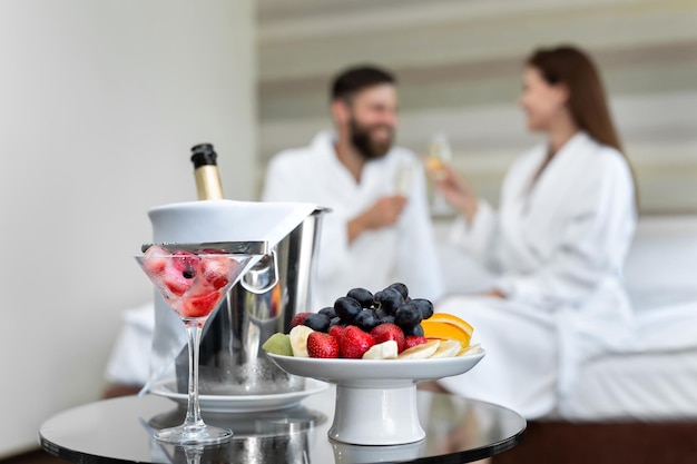 Cena romántica en el hotel de frutos rojos y frutas para una joven pareja que bebe champagne en la cama.